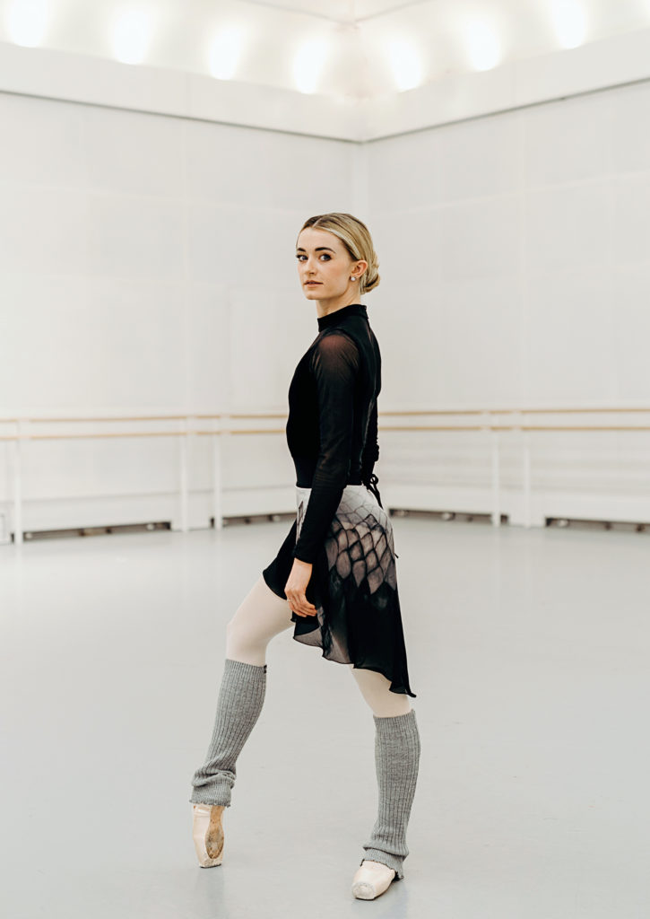 Anna Rose O'Sullivan poses in a large dance studio. She stands in profile facing the left, her arms relaxed at her sides and her right foot propped up onto pointe. She turns her head to look at the camera. She wears a black turtleneck leotard, a black and gray patterned skirt, pink tights, gray legwarmers and pointe shoes.