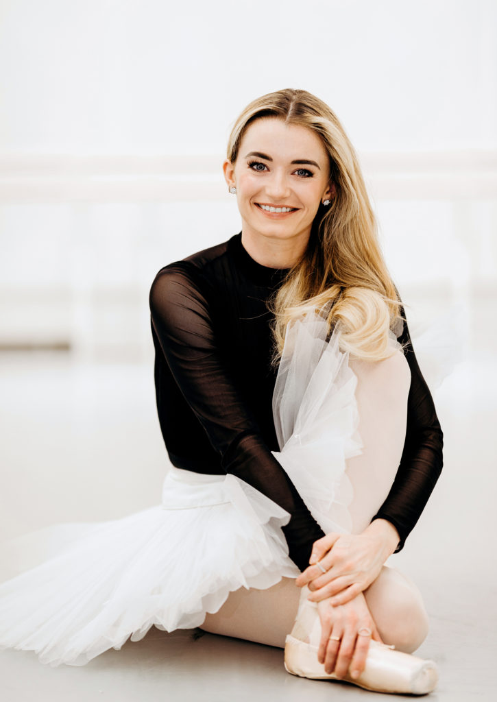 Anna Rose O'Sullivan, her blond hair long, sits on the floor in a dance studio, crossing one leg underneath her and crossing the other one over. She wears a black turtleneck leotard, white practice tutu, pink tights and pointe shoes, and she looks directly at the camera and smiles.