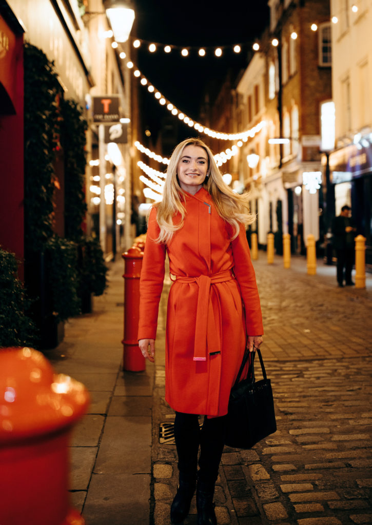 Anna Rose O'Sullivan stands outside on a cobblestone street at nighttime. The streets are decorated with strung white lights, wreaths and other Christmas decor. O'Sullivan wears a bright red raincoat, dark pants and heeled boots, and she carries a large black purse. She smiles towards the camera.