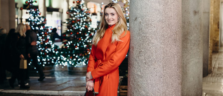 Anna Rose O'Sullivan stands against a pillar in an outdoor square at nighttime. She wears a red raincoat and holds a black purse in her hands, which are resting in front of her. In the background are a row Christmas trees with white and red lights.