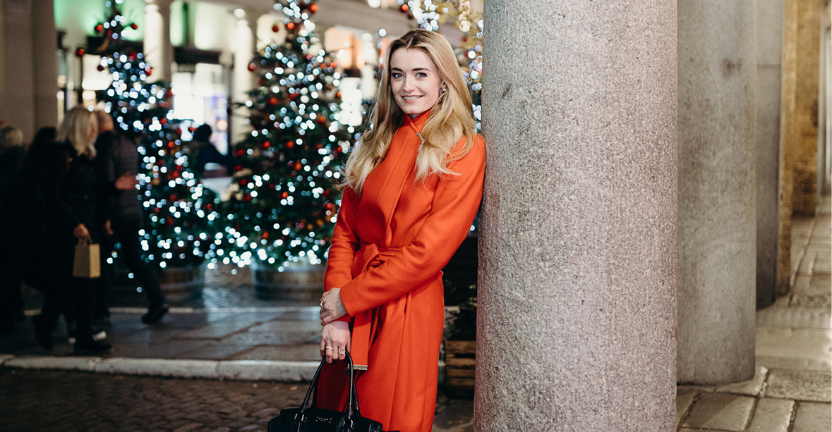 Anna Rose O'Sullivan stands against a pillar in an outdoor square at nighttime. She wears a red raincoat and holds a black purse in her hands, which are resting in front of her. In the background are a row Christmas trees with white and red lights.