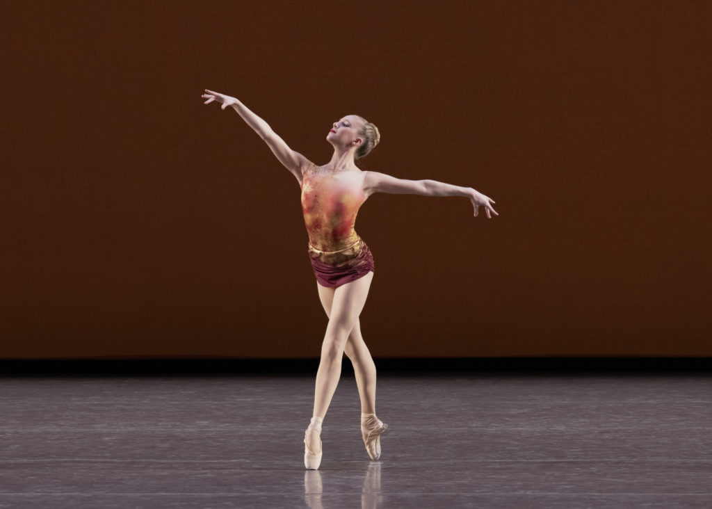 Quinn Starner balances in fourth position en pointe, chin raised smartly to look past her extended arm. Her hair is neatly pulled back in a bun; she wears a leotard-esque costume in oranges and reds over pink tights.