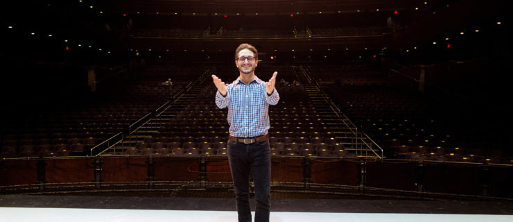 A man in black pants, dress shoes, glasses and a light blue button down shirt stands at the front of the stage, facing away from the audience and toward the camera. He smiles brightly, arms lifted front with his hands open in a motion of gratitude.