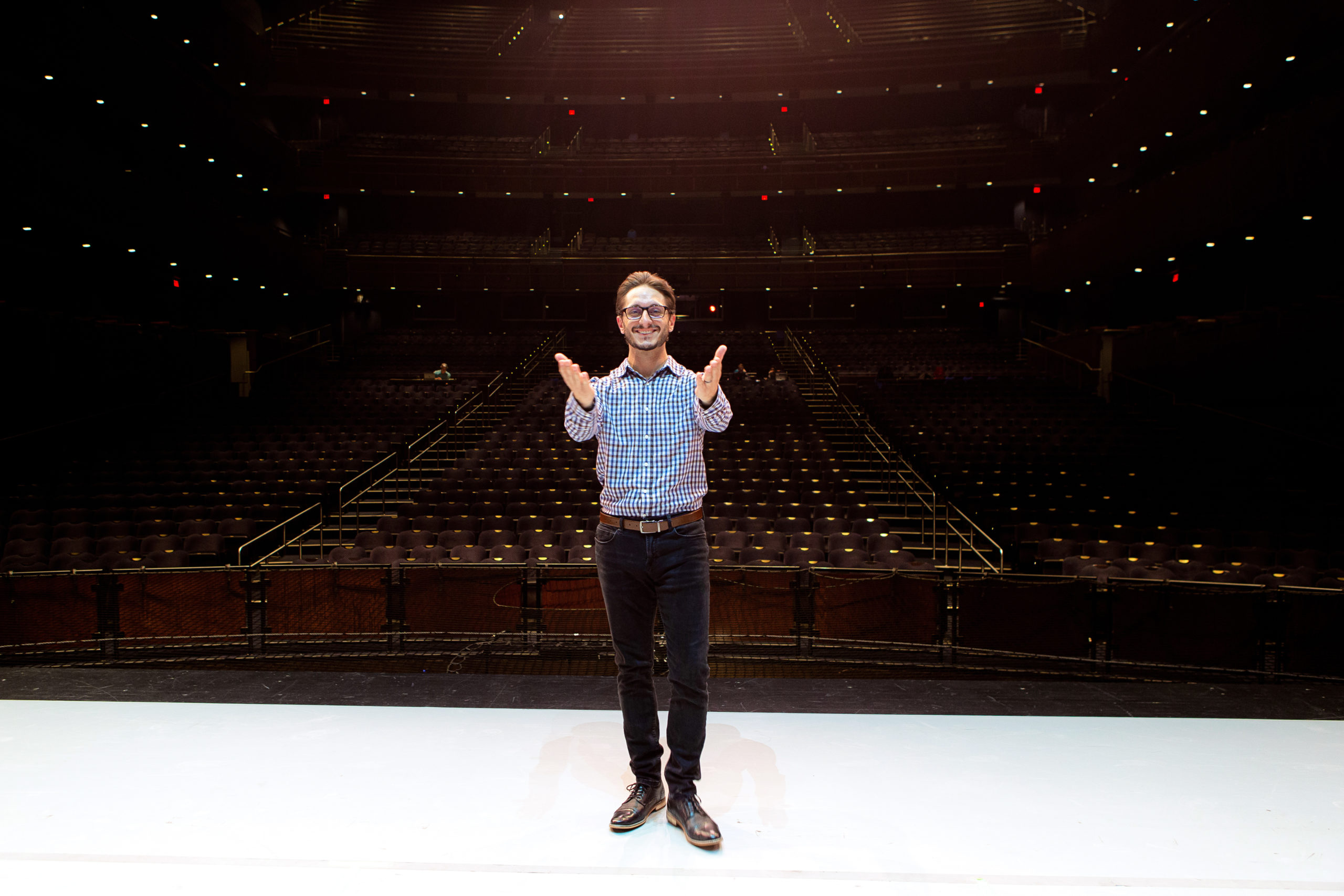 A man in black pants, dress shoes, glasses and a light blue button down shirt stands at the front of the stage, facing away from the audience and toward the camera. He smiles brightly, arms lifted front with his hands open in a motion of gratitude.