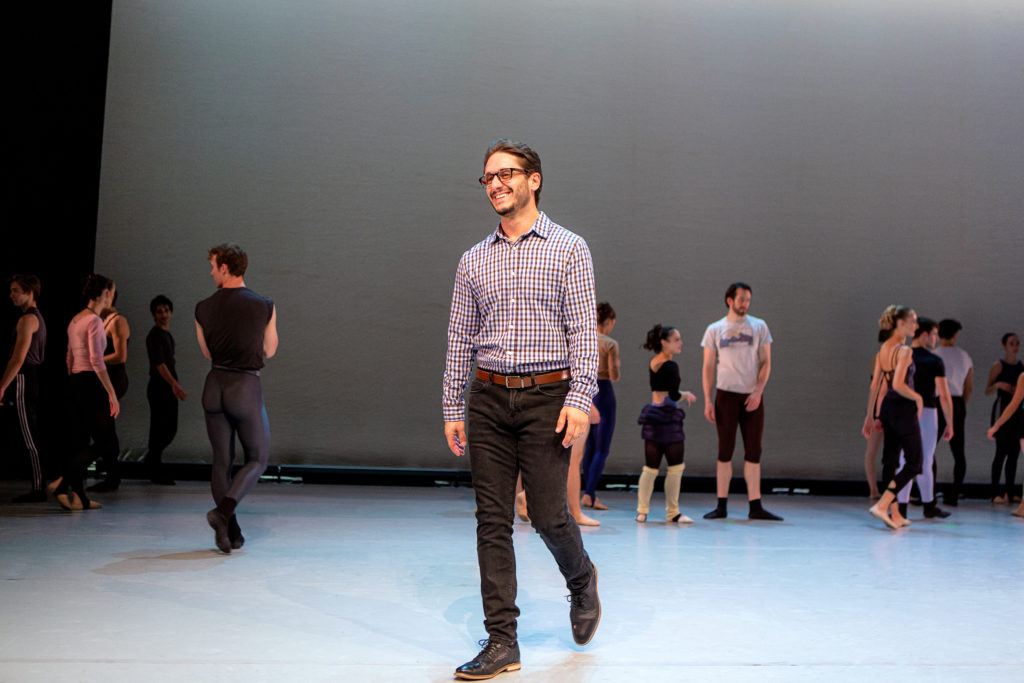 A man in a light blue button down, black pants, dress shoes and glasses walks forward on the diagonal, smiling. Ballet dancers in athletic wear and warmups talk and stand behind him onstage.