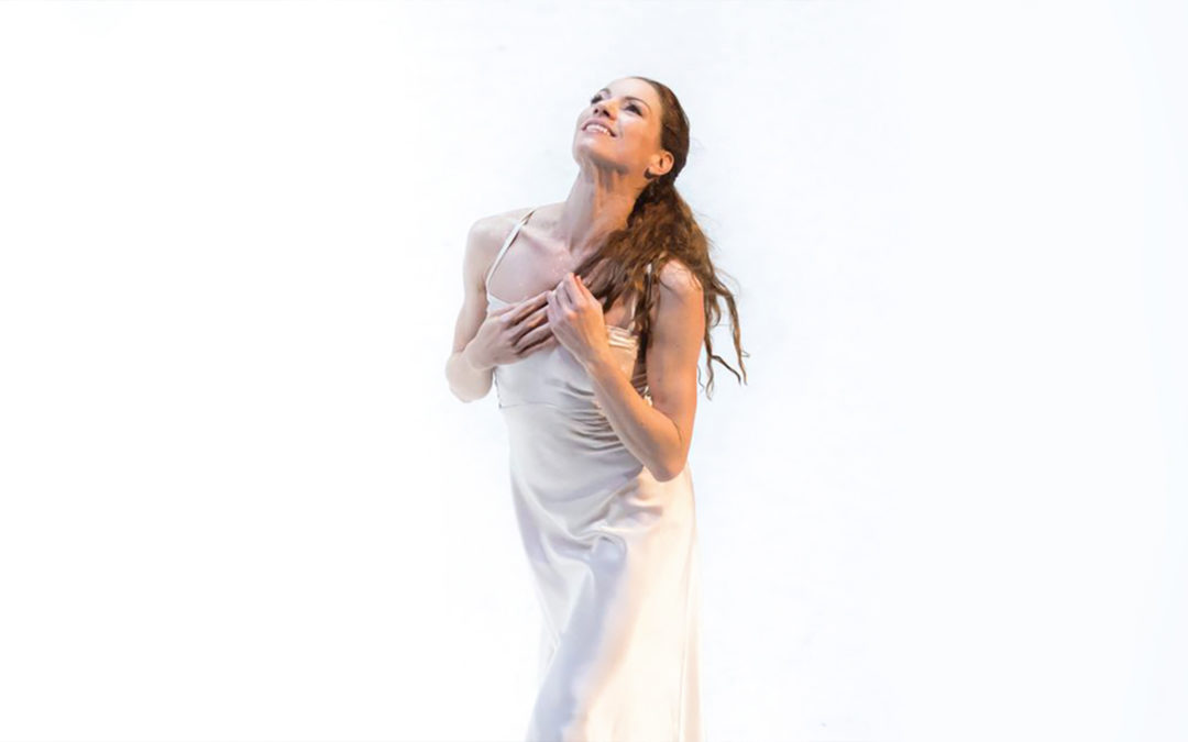 In front of a bright white background, a female ballerina smiles upward with her hands on her chest, playing with her long brown hair. She walks forward and wears a long silky white dress.