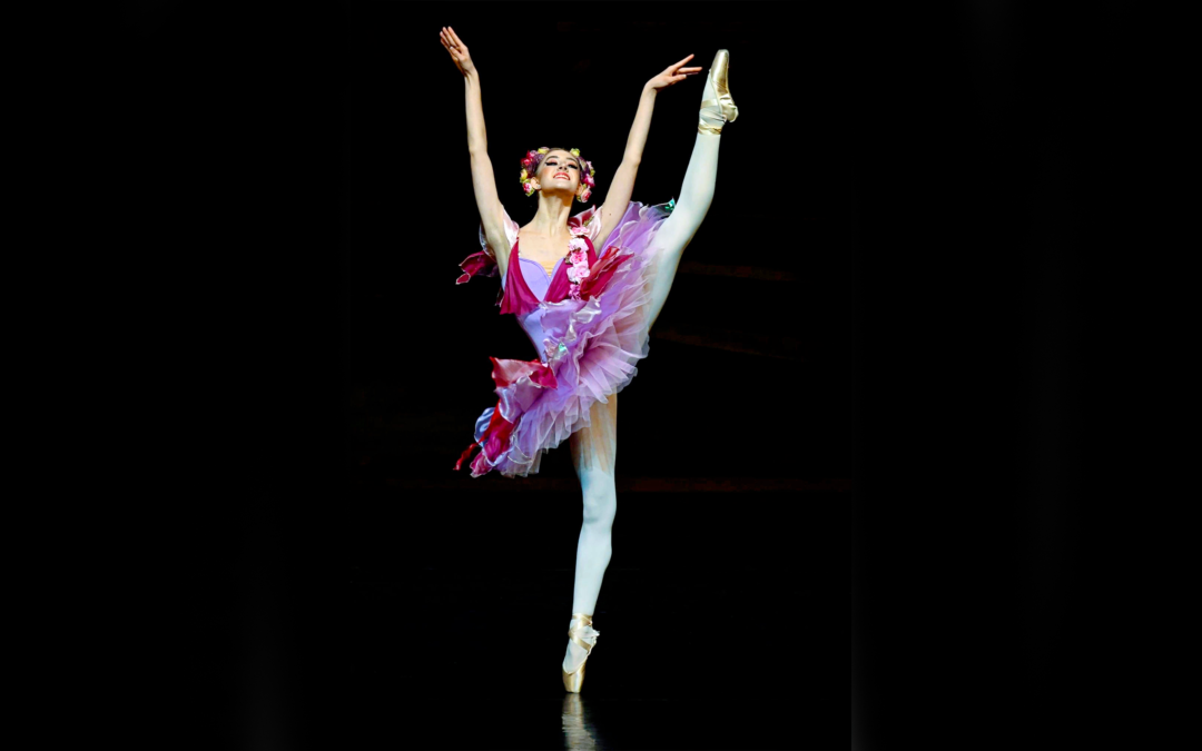 A female ballerina poses on pointe with her left leg in high a la seconde, her arms lifted in a "V" above her head. She wears a floral-themed magenta and lilac-colored tutu with rose adornment and a floral headpiece, with pink ballet tights and pointe shoes.