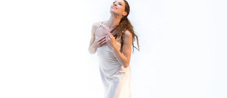 In front of a bright white background, a female ballerina smiles upward with her hands on her chest, playing with her long brown hair. She walks forward and wears a long silky white dress.