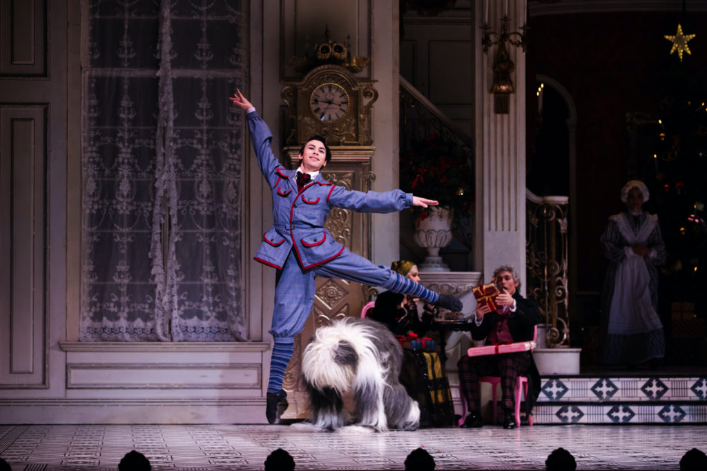 Simone Acri is midair, doing a temps levé. He is costumed in an old-fashioned, childlike blue suit with red piping. A dancer costumed as a shaggy dog appears behind him, seeming ready to pounce.