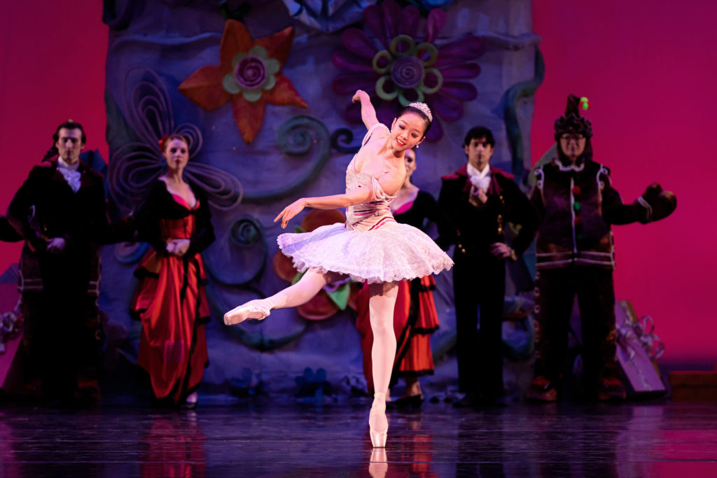 Sirui Liu stands on her left leg on pointe and lifts her right leg in a battement degagé in effacé while crossing her left amr in front of her and her right arm back. She wears a pink tutu, tights and pointe shoes and a tiara. A group of dancers in Spanish-style costumes stand behind her and watch.