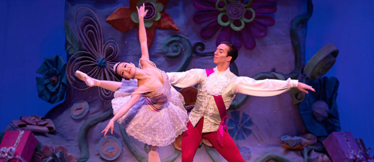 Sirui Liu and Joshua Stayton perform the grand pas de deux from The Nutcracker onstage. Liu poses in an attitude derriere on pointe, leaning off of her right leg and bending her upper body to the right as Stayton holds her waist with his right hand. She ewars a purple tutu, pink tights, pointe shoes and a tiara, while Stayton wears a white top with lacy collar, a red sash, red knee-length knickers and white tights.