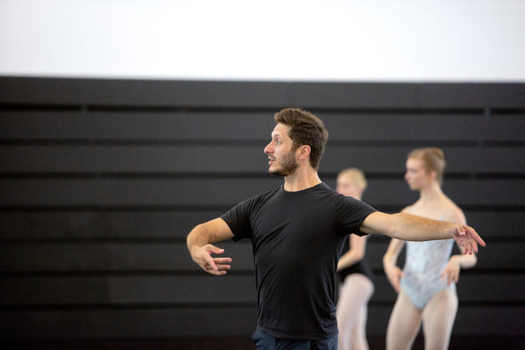 Shown waist-up, a male dance teacher in black athletic clothing demonstrates third port de bras in a studio. Behind him, young ballet students watch in the mirror.
