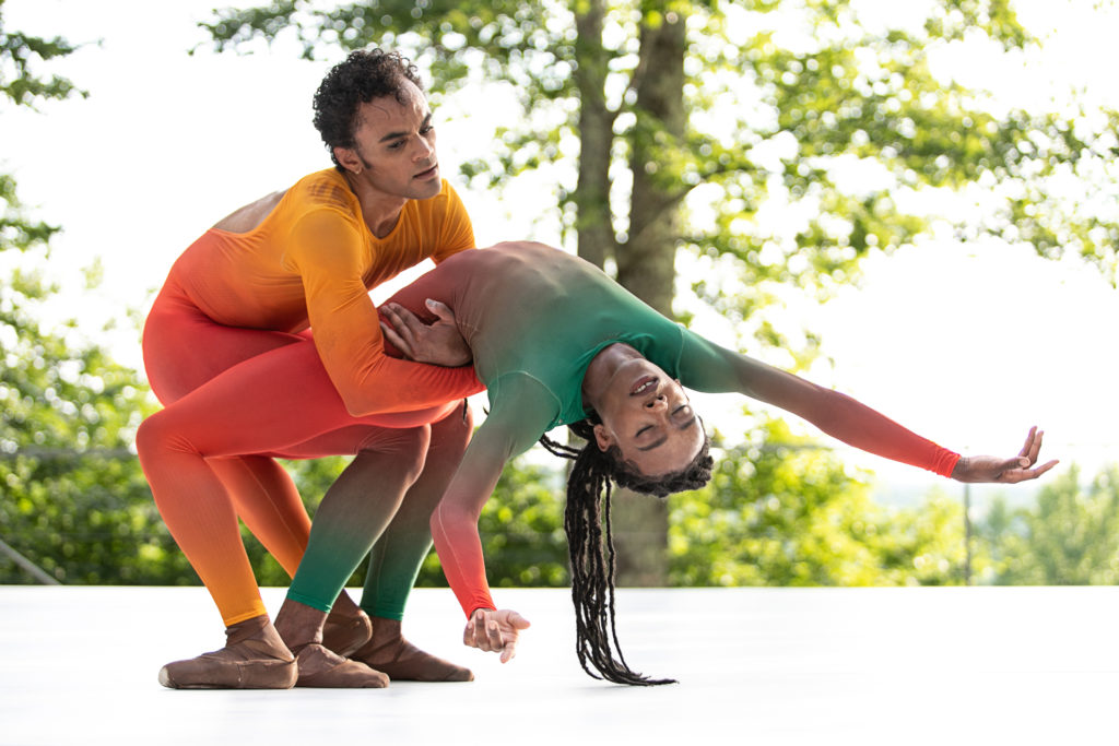 During an outdoor performance, Taylor Stanley, wearing a pink and prgane unitard, crouches low and holds Ashton Edwards by the waist as Edwards does a deep backbend. Edwards wears a green and pink multi-colored unitard and pointe shoes.