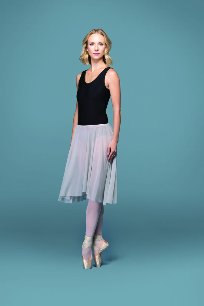 Victoria Hulland stands on pointe in sous-sus in front of a light blueish-gray backdrop, looking at the camera with a slight smile. Her short blonde hair is down and wavy, and she wears a black tank-sleeve leotard, a mid-length gray flowing skirt, pink tights and pointe shoes. Her arms rest gently at her sides.