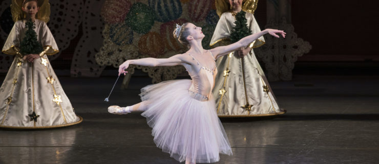 Sterling Hyltin, wearing a pink, knee-length tutu and sparkly tiara, performs the Nutcracker onstage. She is shown doing a piqué arabesque in fourth position on pointe towards stage left, and she holds a wand in her right hand. Behind her, small children costumed as angels hold small evergreen trees in their hands and watch.