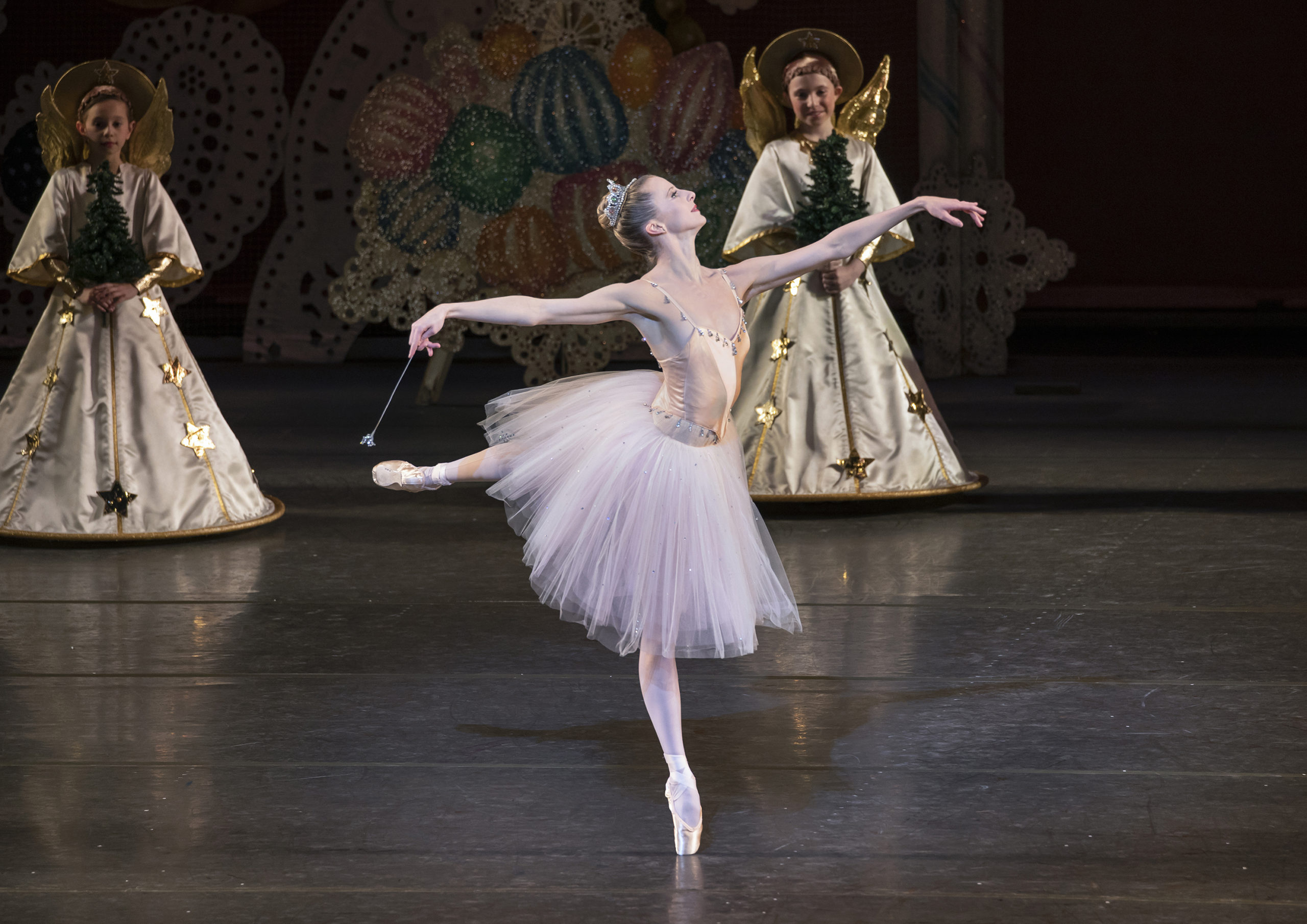 Sterling Hyltin, wearing a pink, knee-length tutu and sparkly tiara, performs the Nutcracker onstage. She is shown doing a piqué arabesque in fourth position on pointe towards stage left, and she holds a wand in her right hand. Behind her, small children costumed as angels hold small evergreen trees in their hands and watch.