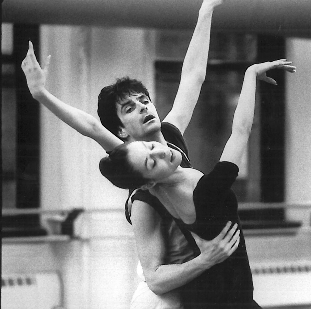 In this black and white photo, Kevin McKenzie and Susan Jaffe are shown from the waist up partnering together in a rehearsal in a dance studio. McKenzie stands behind Jaffe and holds the right side of her rib cage as she leans back with her arms in third arabesque.