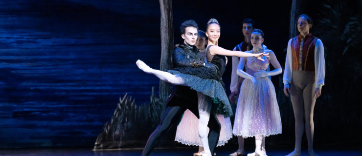 Hui Wen Peng and Carraig New perform "Swan Lake" onstage. Peng wears a black tutu embellised with black feathers, a tiara, pink tights and pointe shoes and poses in attitude effacé with her right leg back and her right arm across the front of her body. Cruz Alvarez stands behind her, dressed all in black and with dramatic makeup on, and holds her waist. Behind them, other dancers in costumes watch.