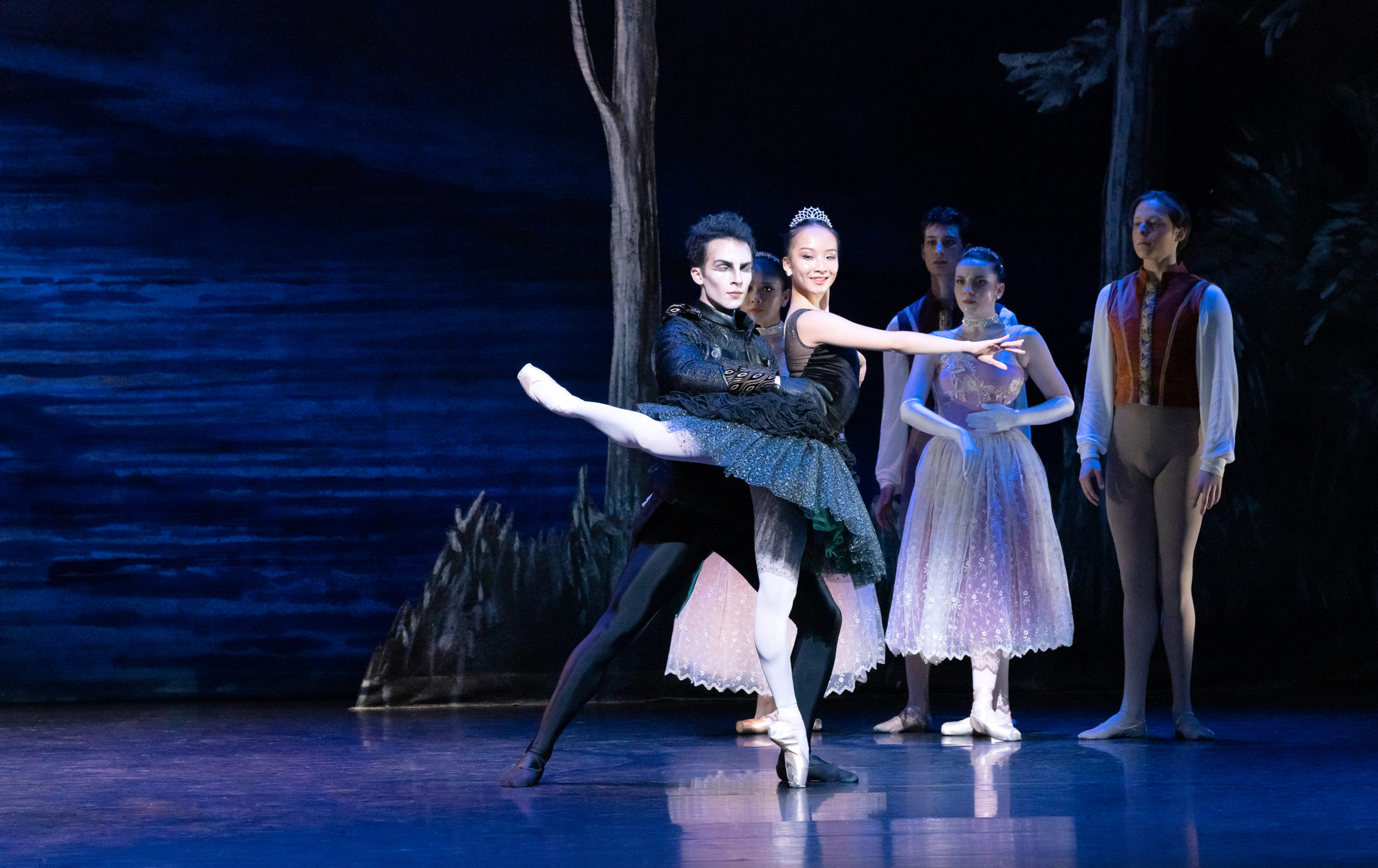 Hui Wen Peng and Carraig New perform "Swan Lake" onstage. Peng wears a black tutu embellised with black feathers, a tiara, pink tights and pointe shoes and poses in attitude effacé with her right leg back and her right arm across the front of her body. Cruz Alvarez stands behind her, dressed all in black and with dramatic makeup on, and holds her waist. Behind them, other dancers in costumes watch.