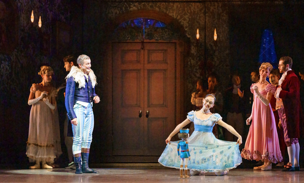 During a stage performance of The Nutcracker, a smiling young dancer in a light blue floral dress kneels on the floor in front of a Nutcracker doll and stretches her skirt to the sides.  To his left is an older male figure dressed in a blue jacket with a fur collar, white and blue striped pants, and dark boots.  He looks at her and smiles as other dancers in Regency period costumes stand behind them and watch.