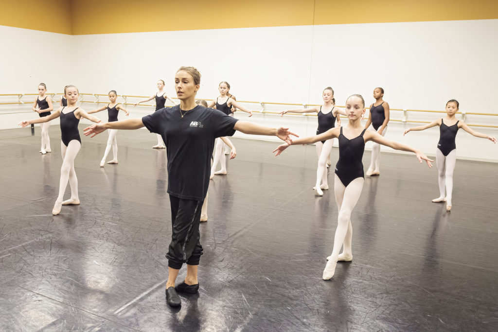 A ballet teacher leads a group of young girls in a tendu exercise.