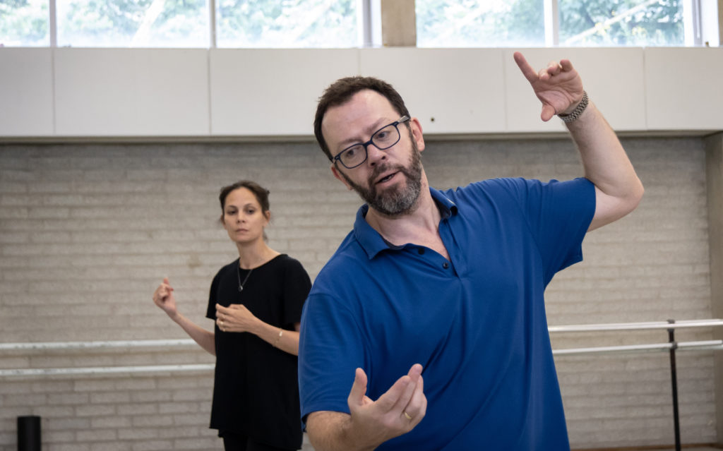 Alexei Ratmansky, shown waist-up, wears a blue polo shirt as he demonstrates a movement with his words and upper body during rehearsal. A female dancer in a black shift stands behind him and watches.