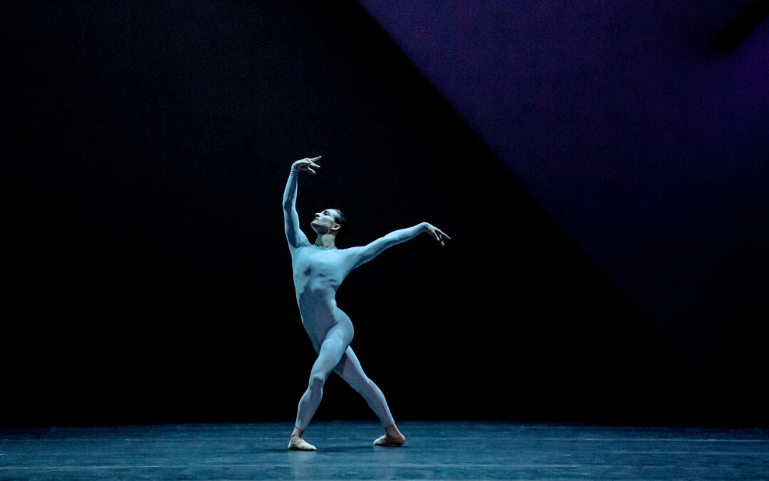Bathed in blue light, Shale Wagman performs onstage wearing a blue unitard. He poses in a large fourth position croisé with his left leg in front. He raises his curved right arm up and looks toward his hand and reaches his left arm to the side, slightly higher than his shoulder.