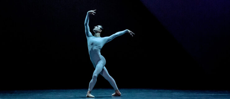 Bathed in blue light, Shale Wagman performs onstage wearing a blue unitard. He poses in a large fourth position croisé with his left leg in front. He raises his curved right arm up and looks toward his hand and reaches his left arm to the side, slightly higher than his shoulder.