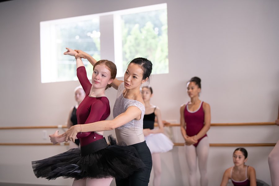 Xuan Cheng stands behind a teenage ballet student and corrects her arms while other dancers look on in the back of the studio. Cheng wears a white leotard and black pants, while her student wears a burgundy long-sleeved leotard, black practice tutu and pink tights.