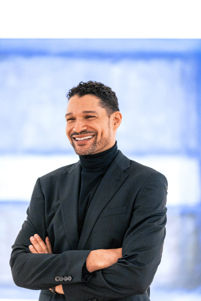 Adam McKinney is shown in three-quarter view, from the waist up, with his arms folded and a big smile.  He is wearing a black turtleneck and black blazer and is standing in front of a blue background.