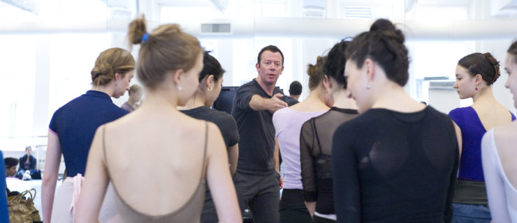 In a brightly lit studio, Alexei Ratmansky stands in front of a mirror, motioning with his hand and speaking to the crowd of female and male dancers in front of him. They wear various shades and styles of dancewear and athletic clothing.
