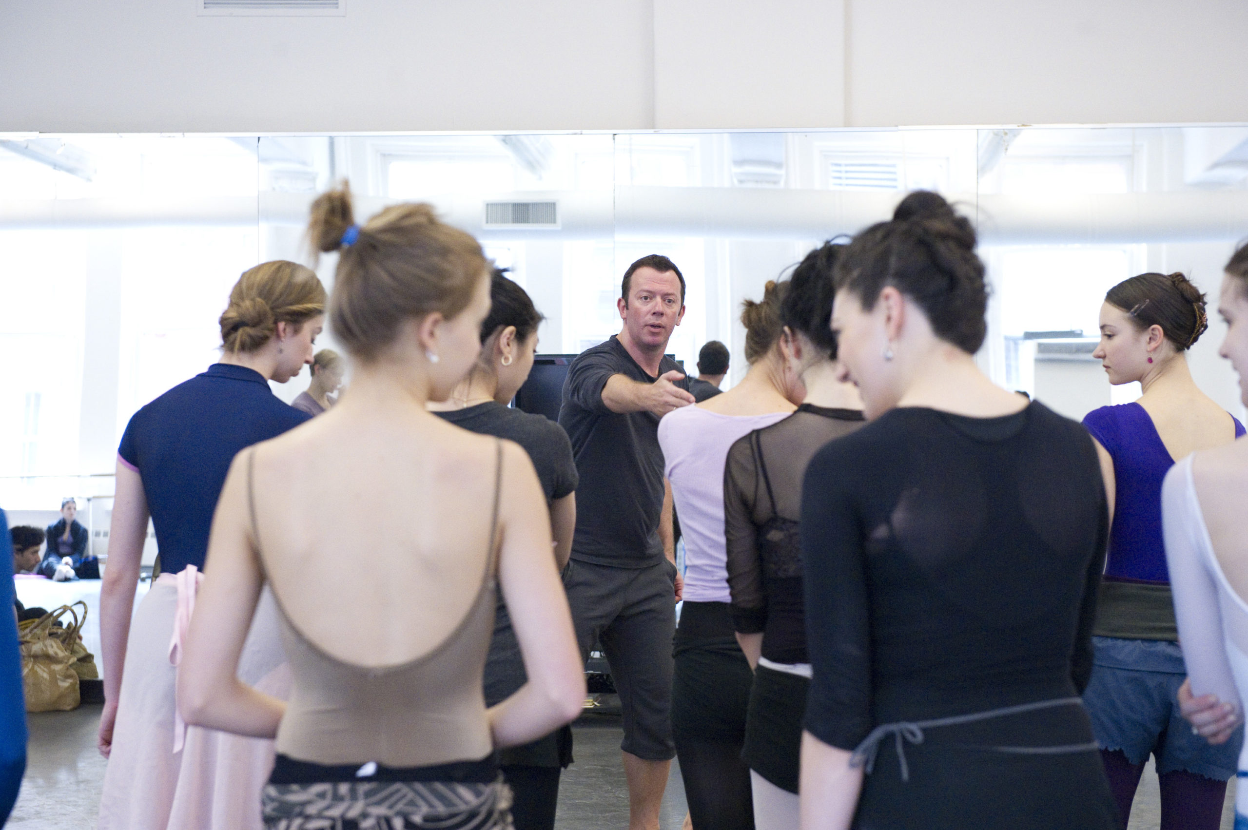 In a brightly lit studio, Alexei Ratmansky stands in front of a mirror, motioning with his hand and speaking to the crowd of female and male dancers in front of him. They wear various shades and styles of dancewear and athletic clothing.