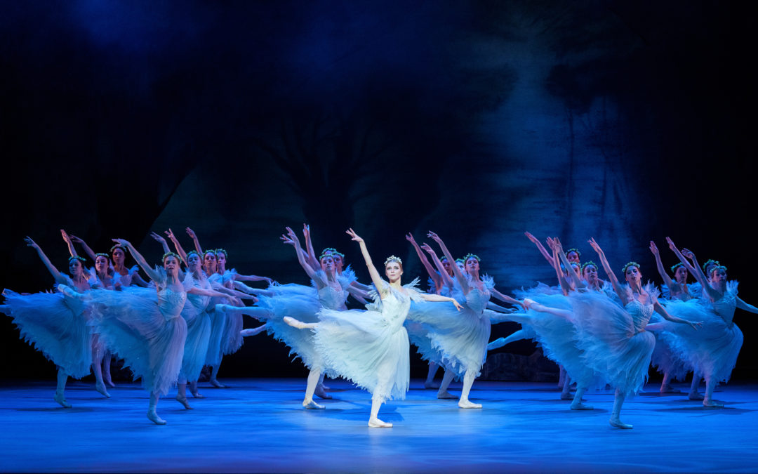 Onstage against a black and dark blue backdrop, with deep blue lighting, a corps de ballet of willis and their leader, Myrtha, pose in an arabesque fondu with arms extended in allongé. They wear long, wispy Romantic tutus and flower crown headpieces.