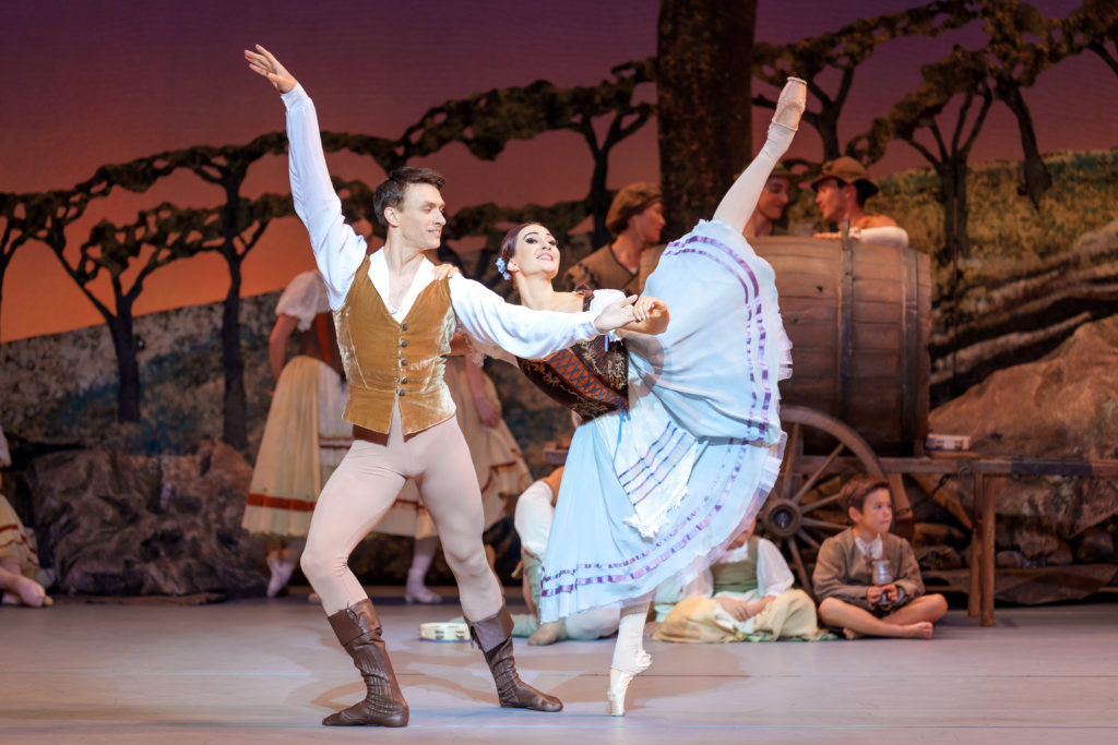 Onstage against a bohemian village backdrop, a female dancer performs as Giselle in a light blue and brown peasant dress. She executes a penché arabesque on pointe, hands on her partner's shoulder and hand for support. Her partner, a male dancer performing as Albrecht, lunges in 4th with his other arm extended and wears a tan and brown peasant costume.