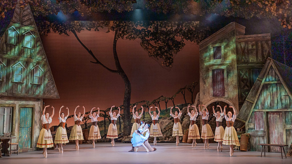Onstage against a backdrop of a charming bohemian village, a corps de ballet of female peasants pose in sous-sus in a semi circle around Giselle and Albrecht. Giselle, in a blue peasant dress, sits on Albrecht's knee as he kneels, and they smile into each others's eyes lovingly.