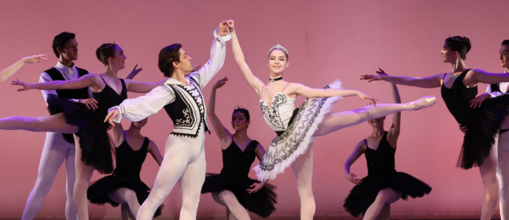 A performance photo of a young male holding a young woman's hand as she balances in arabesque on pointe.
