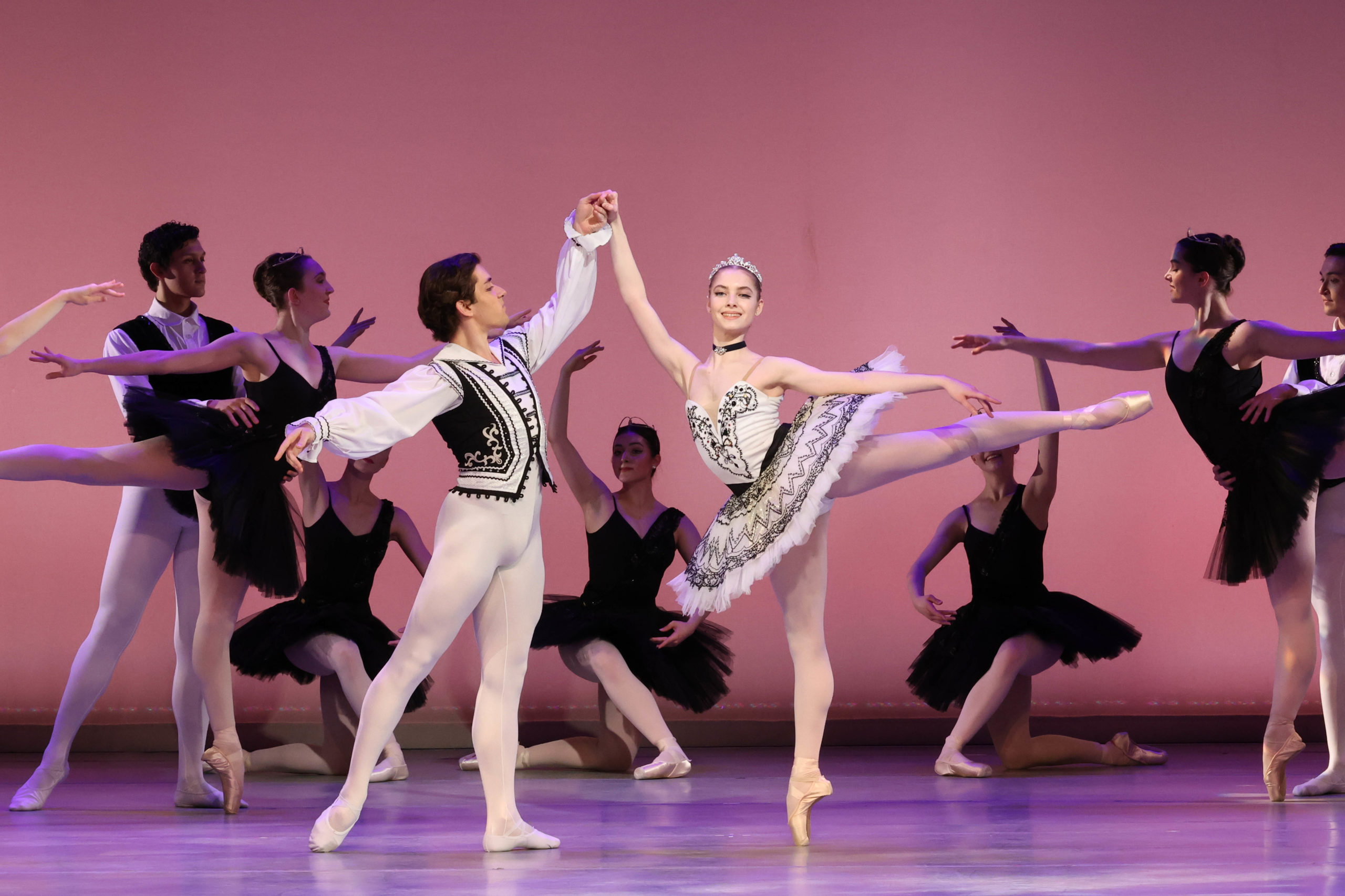 A performance photo of a young male holding a young woman's hand as she balances in arabesque on pointe.