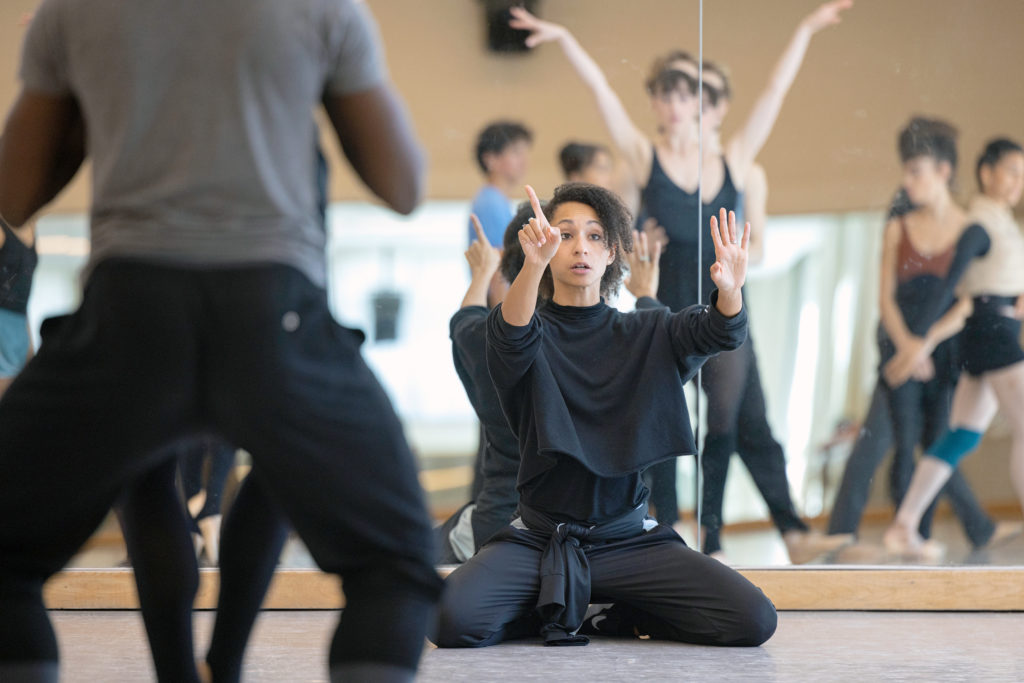 In a brightly lit studio, Claudia Shreier sits on her heels, back against the mirror as she directs her rehearsal. She holds both hands up, pointing with her second finger on her right hand, and speaks to the dancers, who are shown in the reflection of the mirror.