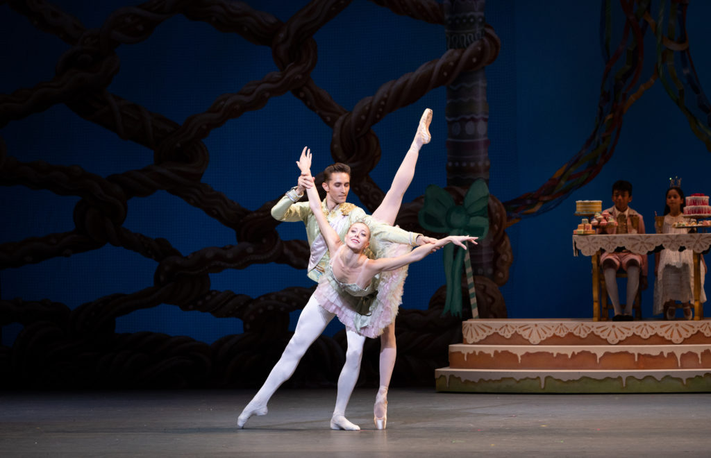 Onstage in front of a navy blue background and red and gold set pieces, Dawn Atkins and Stanislav Olshanskyi perform the Nutcracker grand pas de deux. Atkins wears a sparkling light green and pink pancake tutu with a gold crown, pink tights and pointe shoes. She penches in arabesque towards the audience, arms spread wide in allonge as Olshanskyi supports her holding on to her wrists. He lunges in a tendu a la seconde and wears a light green, gold-embellished jacket with a pink sash, pink tights and ballet shoes. 