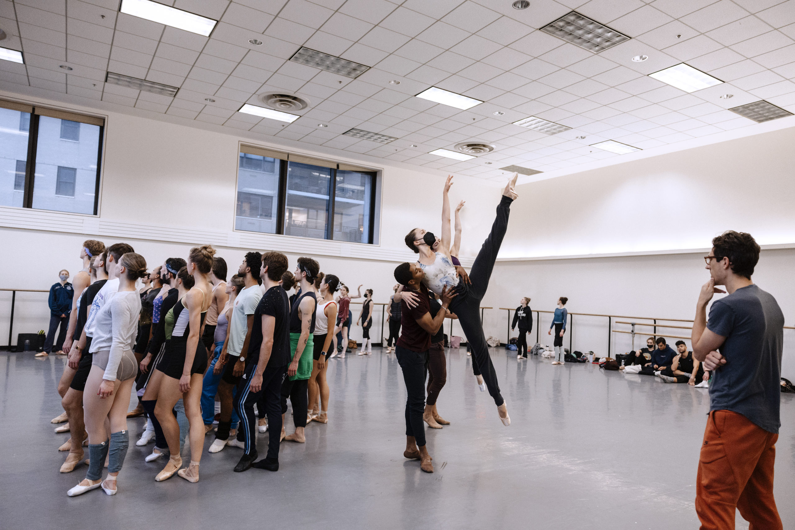 In a large dance studio, a big group of male and female dancers stand in a clump toward the middle of the studio. To the left of them, two pairs of male and female partners perform a supported lift, with the women leaning against their partners in an airborne a la seconde position. Justin Peck, in a red shirt and black pants, stands to the far right and watches the dancers.