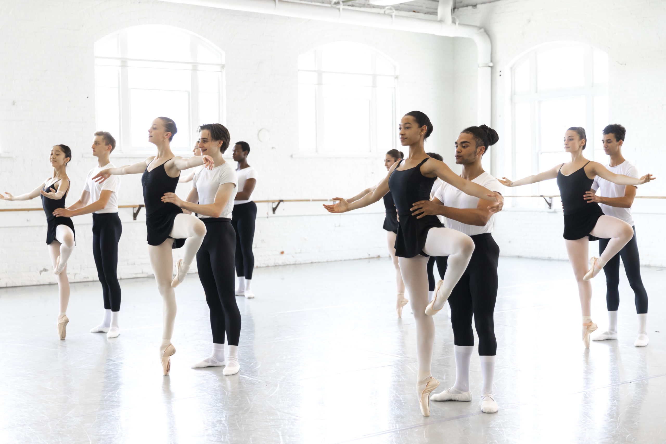 Six teen ballet couples in a studio. The women balance in passé relevé as the men support them, holding their waists from behind.