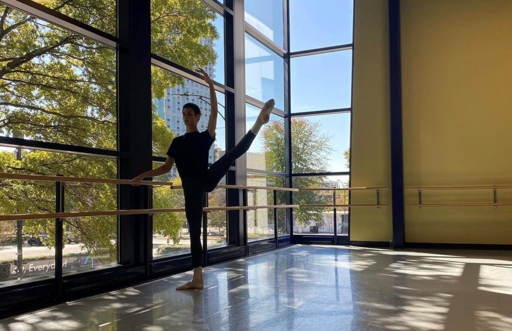 A young male dancer does a développé to the side at the barre in a studio with a wall of windows.