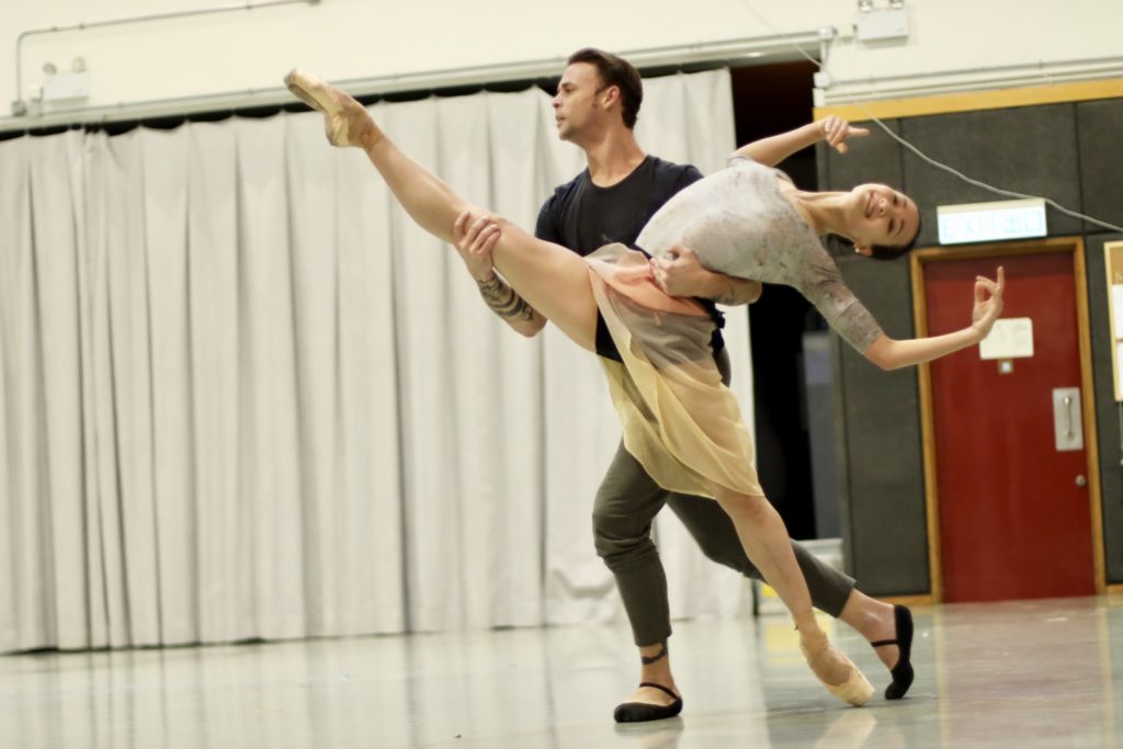 Two dancers rehearse a passionate pas de deux in a large dance studio. The woman rises onto pointe and lifts her right leg high and leans back into her male partner's arms. He holds her by the waist and her right leg leg and runs across the room, sliding her along in a split. She warsa a gray leotard and cream-colored skirt, while he wears a black T-shirt and gray tights.