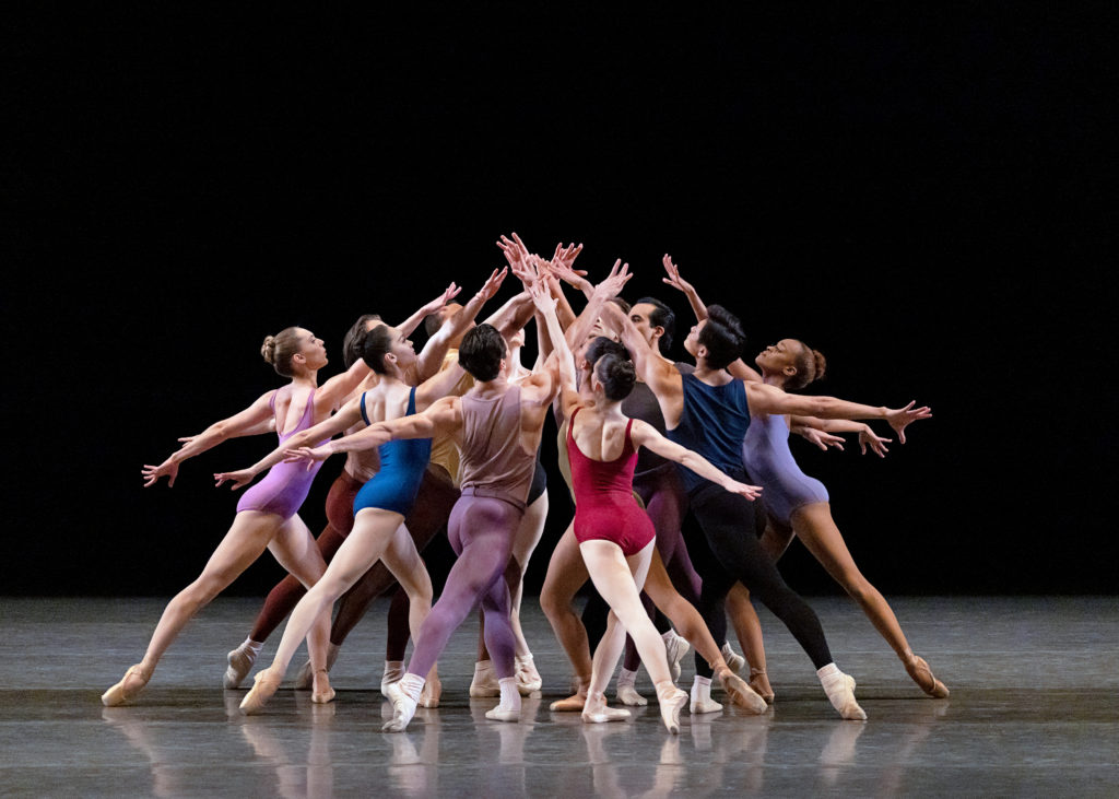 During a performance, a diverse group of male and female dancers form a circle and lunge towards each other with their back legs in tendu. The women wear leotards in various colors and skin-tone tights and pointe shoes. The men wear tank tops and tops in various colors, as well.