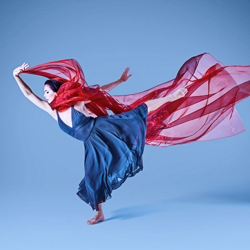 In a long, silky blue dress, Natalia Osipova poses in fondu arabesque in front of a light blue backdrop. Her arms in a free first arabesque line, she holds a long, flowing piece of transparent red fabric in her right hand above her head. The fabric trails behind her, extending the line of her left leg in arabesque.