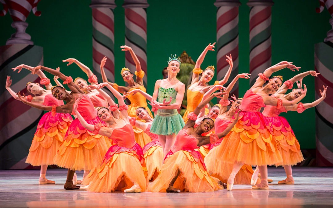 During a performances of The Nutcracker, a lead ballerina stands in B plus encircled by a corps de ballet of dancers in long, orange and pink tutus. Four of the dancers kneel on the ground, while the others stand in tendu devant croisé. All hold their arms in high fifth and lean their upper bodies out away from the circle. The soloist wears a short green dance dress and crown and crosses her arms in front of her with her palms up.