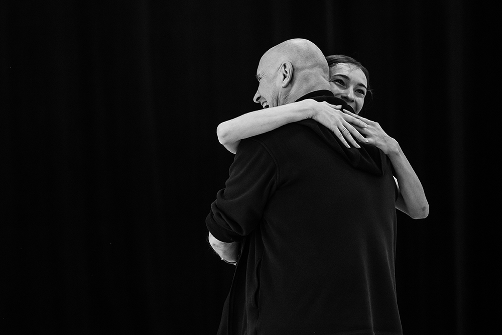 A black and white photo shows Olga Smirnova and Jean-Christophe smiling and hugging in front of a black background.
