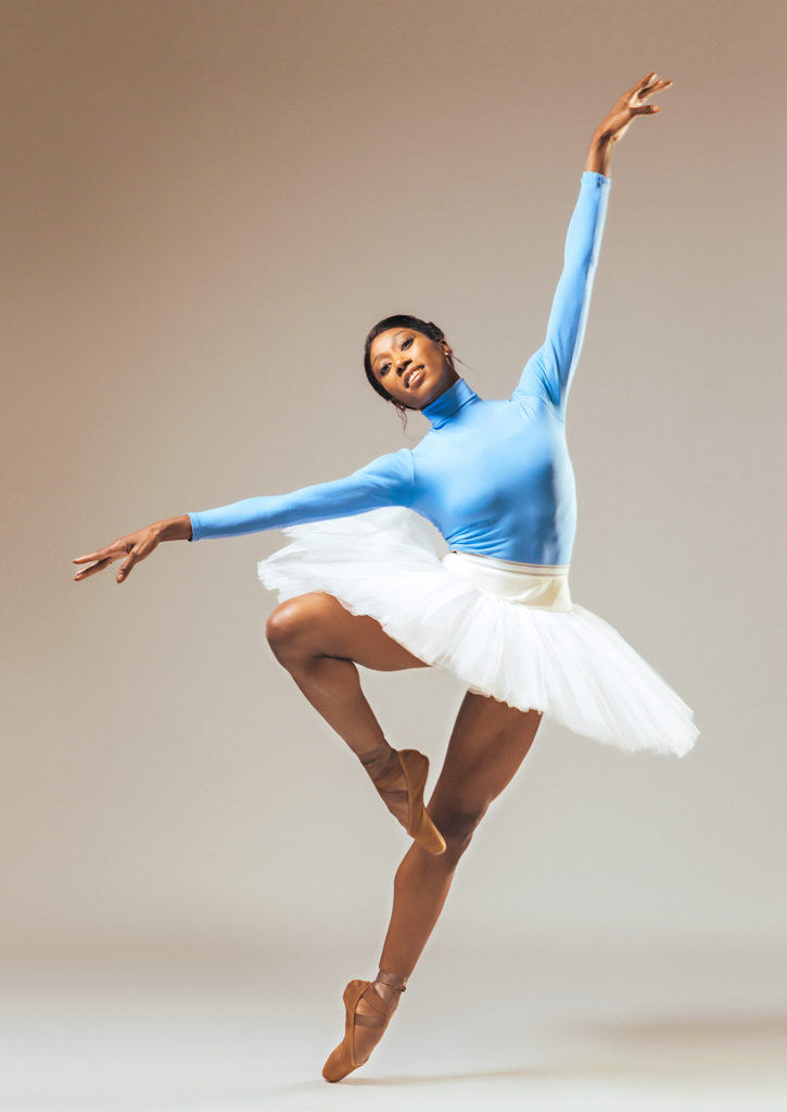 Claudia Monja does a passé relevé on pointe and leans into her left hip, tilting slightly off balance on her left foot. She opens her arms out, with her left arm lifted and her right arm lowered on a slight diagonal, and smiles peacefully. She wears a blue turtleneck leotard, white practice tutu and brown pointe shoes and dances in front of a gray backdrop.