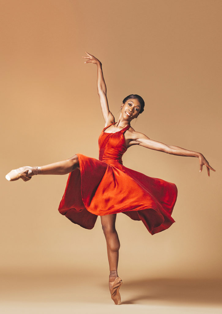 Claudia Monja, wearing a silky, flowing red dress, poses in an attitude devant on pointe. She opens her arms wide, her right arm up high and her left out to the side. She leans her face slightly to the left and smiles, her skirt swirling out around her.