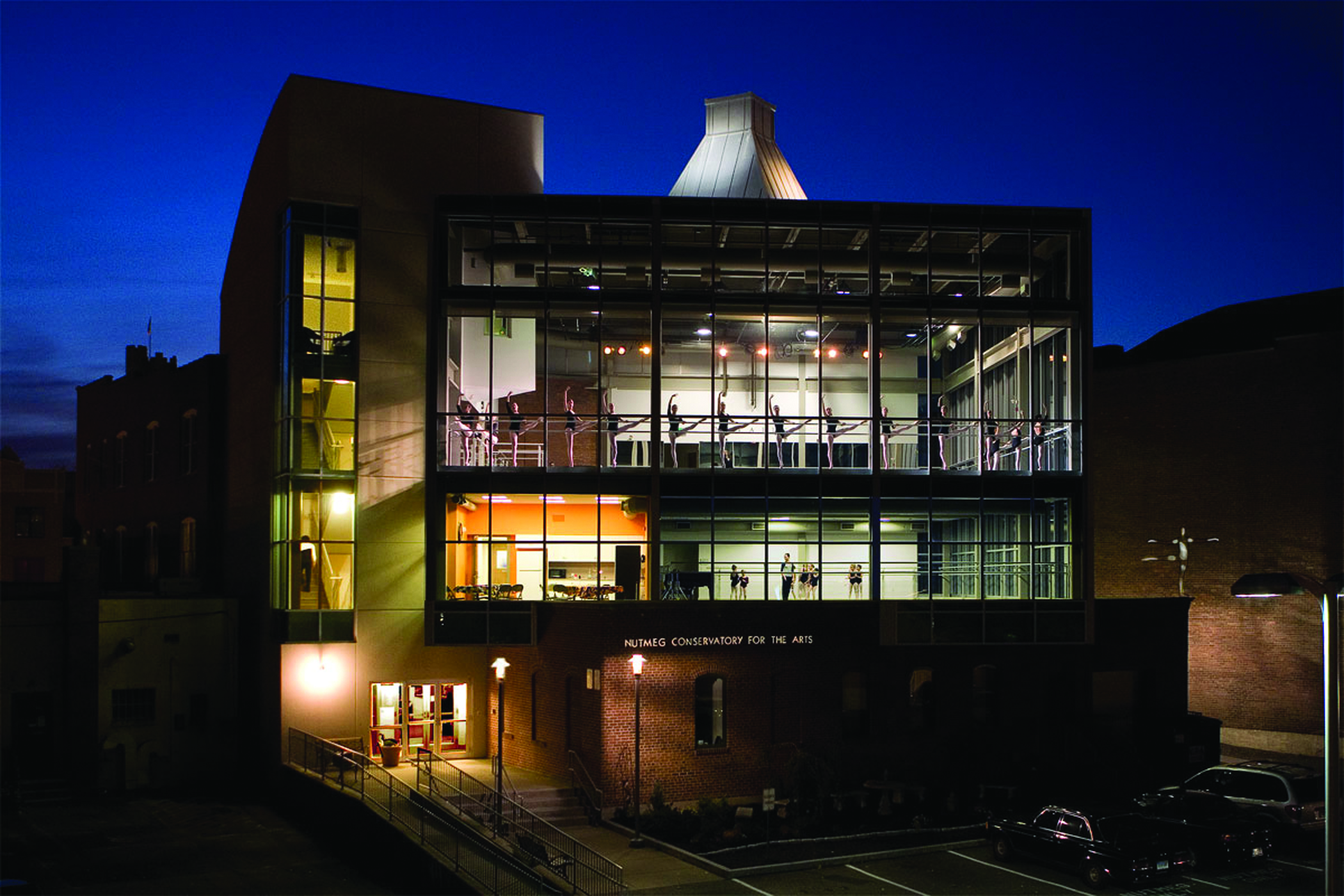 Nutmeg Conservatory's dance studios seen from outside at night. Ballet dancers are seen through the windows in two studios across two floors.